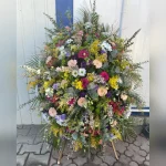 Pełen kolorów wieniec z gerber, margaretek i róż, otoczony zielenią liści palmy. Podkreśla piękno wspomnień i czułość pożegnania.