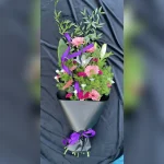 Elegancki bukiet kondolencyjny z różowej gerbery, goździków i lilii, ozdobiony ruskusem, asparagusem oraz fioletową wstążką, otoczony czarną bibułą.