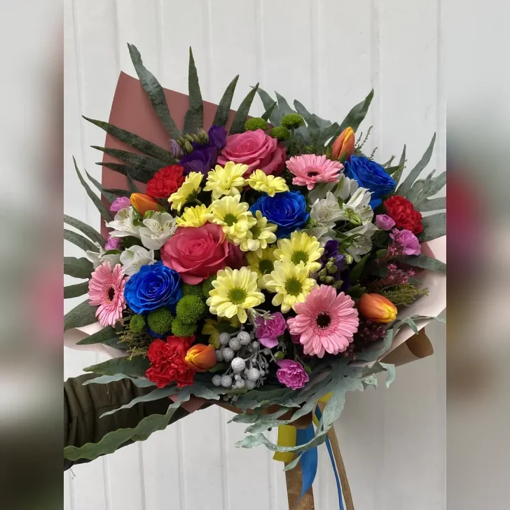 Barwny bukiet z margaretek, gerber, eustom, alstromerii, goździków i niebieskich róż. Zieleń paproci i neutralna bibuła podkreślają intensywność kolorów.