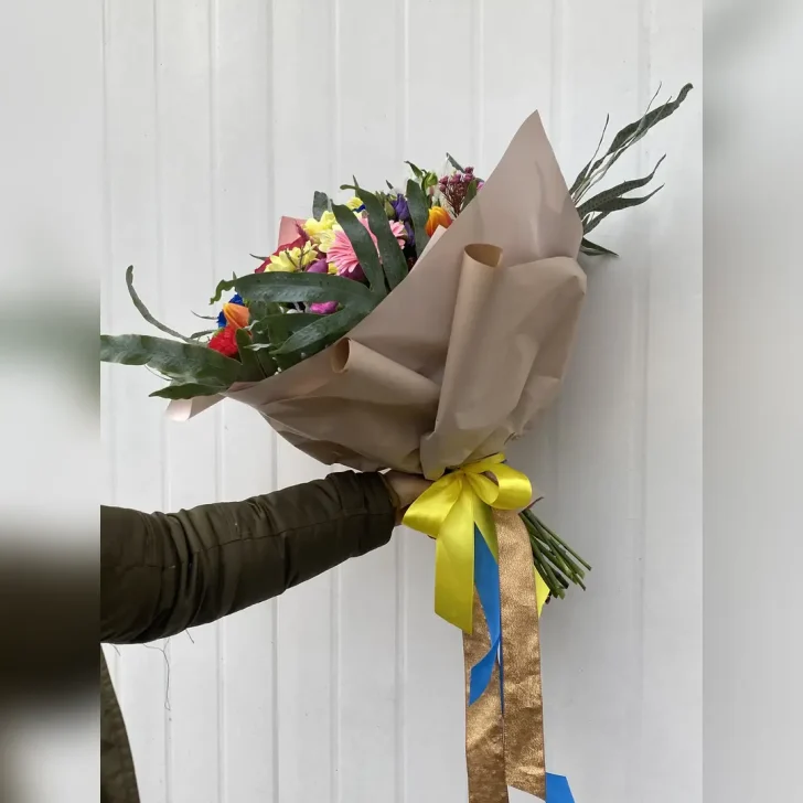 Barwny bukiet z margaretek, gerber, eustom, alstromerii, goździków i niebieskich róż. Zieleń paproci i neutralna bibuła podkreślają intensywność kolorów.