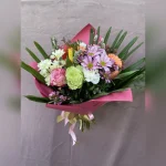 Kolorowy, radosny bukiet z róż, margaretek, goździków i tulipanów, ozdobiony baziami. Różowa bibuła i złoto-różowa wstążka dodają mu uroku.