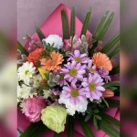 Kolorowy, radosny bukiet z róż, margaretek, goździków i tulipanów, ozdobiony baziami. Różowa bibuła i złoto-różowa wstążka dodają mu uroku.