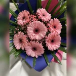 Subtelny bukiet z dużych, jasnoróżowych gerber i gipsówki, podkreślony ciemnozielonymi liśćmi. Całość otacza elegancka granatowo-różowa bibuła, nadając mu wyjątkowy charakter.