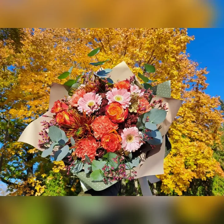 Okrągły bukiet z Kwiaciarni Klara, z pomarańczowych róż, margaretek i goździków, ozdobiony pastelowymi dodatkami oraz papierem w kolorze złotym i szaro-zielonym.