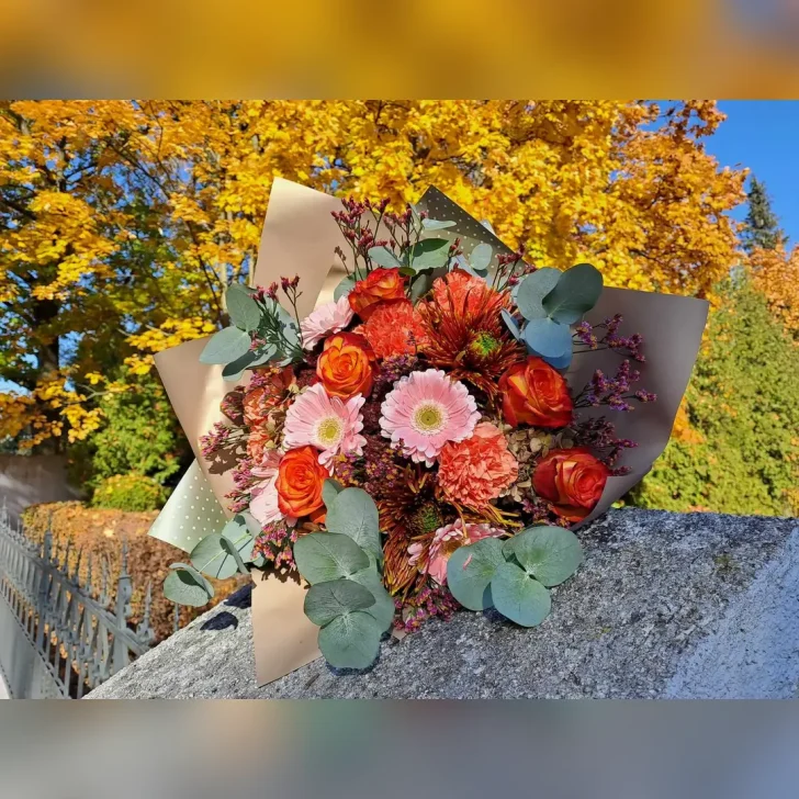 Okrągły bukiet z Kwiaciarni Klara, z pomarańczowych róż, margaretek i goździków, ozdobiony pastelowymi dodatkami oraz papierem w kolorze złotym i szaro-zielonym.