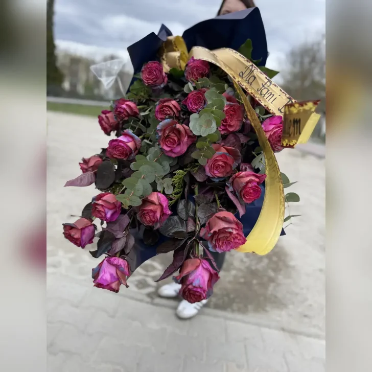 Leżąca pogrzebowa wiązanka z szaro-czerwonych róż, ciemnych bordowych liści i eukaliptusa, oprawiona w czarną folię z szeroką złotą wstążką.