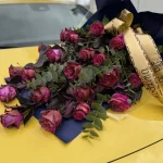 Leżąca pogrzebowa wiązanka z szaro-czerwonych róż, ciemnych bordowych liści i eukaliptusa, oprawiona w czarną folię z szeroką złotą wstążką.