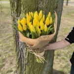 Osoba trzymająca bukiet żółtych tulipanów owinięty zieloną siatką i przewiązany jasnozieloną wstążką – elegancka, minimalistyczna kompozycja kwiatowa.