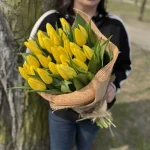 Osoba trzymająca bukiet żółtych tulipanów owinięty zieloną siatką i przewiązany jasnozieloną wstążką – elegancka, minimalistyczna kompozycja kwiatowa.