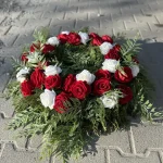 Leżący wianek z żywej tuji, ozdobiony białymi i czerwonymi sztucznymi różami oraz zielonymi liśćmi paproci – dekoracja na płytę nagrobną.