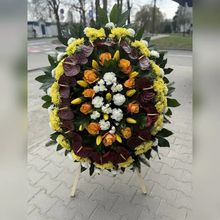 Owalny wieniec z białych goździków, żółtych lilii i tulipanów, z akcentami bordowego anturium i pomarańczowych róż – delikatna i elegancka kompozycja.